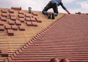 A roofer carefully installs shingles on a new residential roof under a cloudy sky.